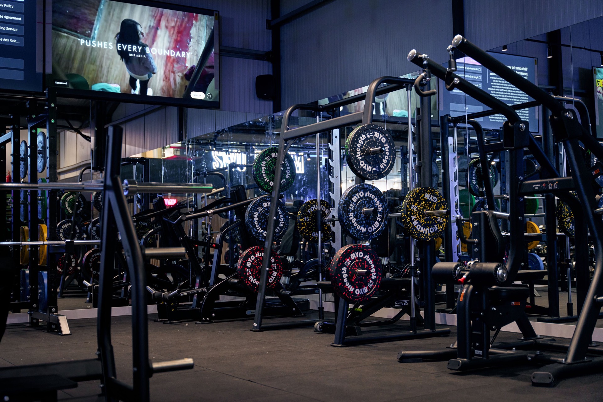 The Swole Gym training floor — black steel racks and chrome under low ambient light, with the neon sign reflected in the mirrors